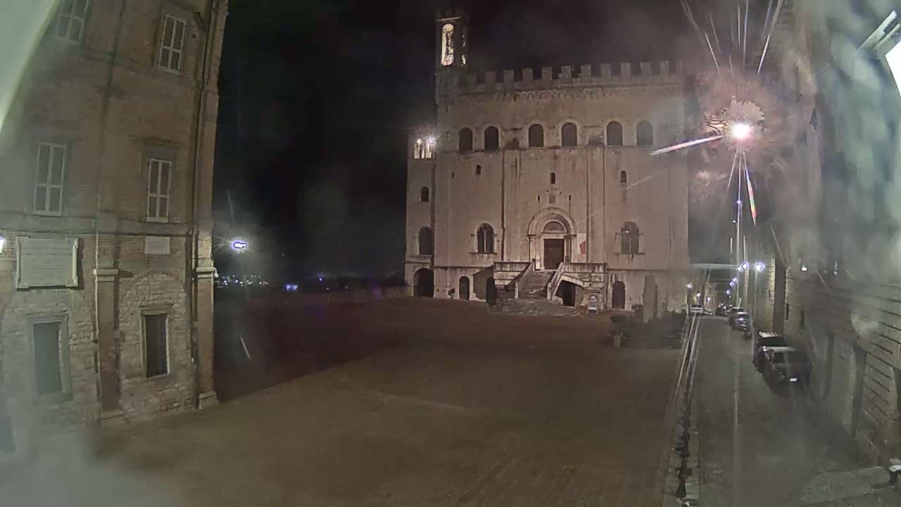 A large, stone building with a staircase stands in a town square at night, with fireworks exploding overhead.
