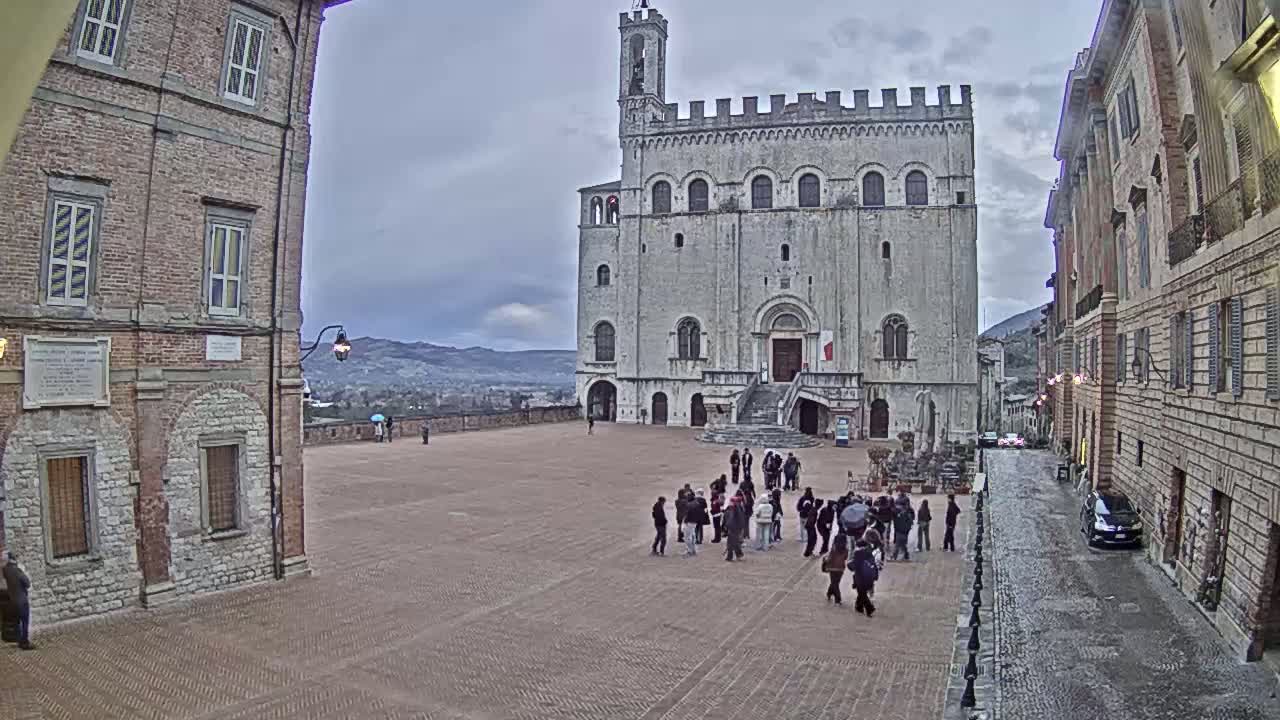 A large, stone building with a staircase stands in a town square, under an overcast sky, with several people gathered in the square and cars parked on a nearby street.