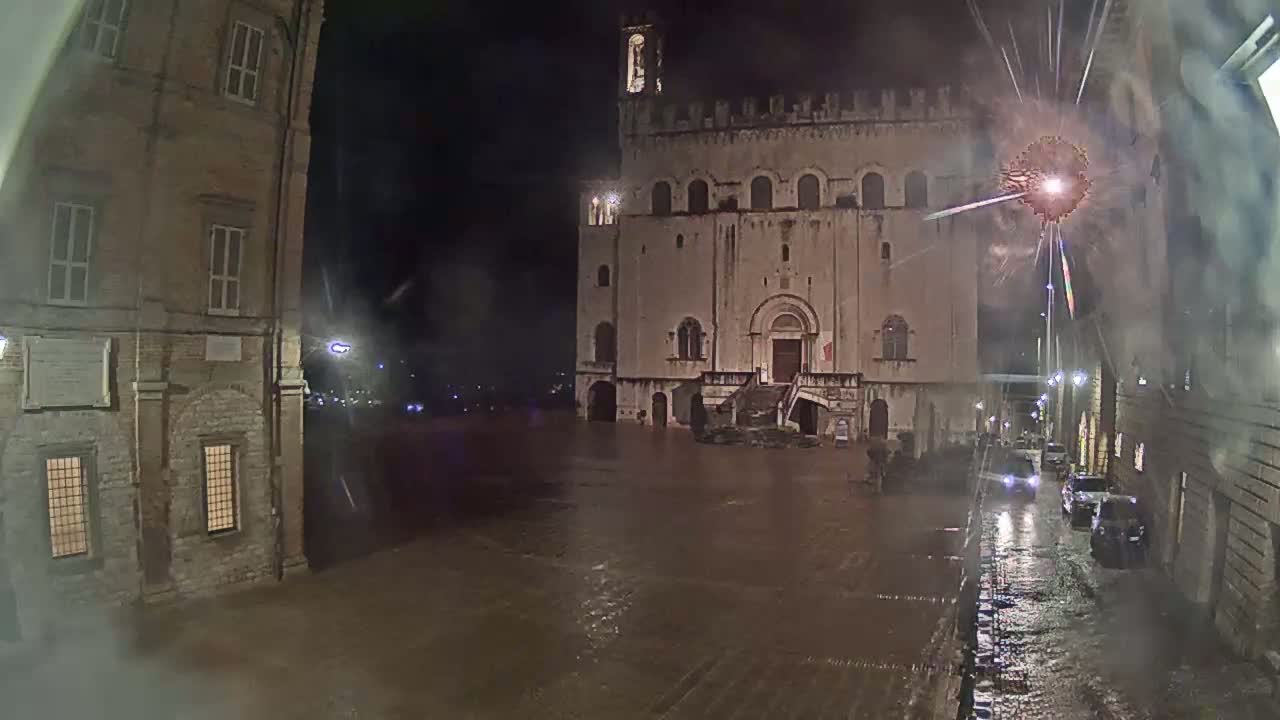 A flooded town square at night, with fireworks exploding over a large stone building, and cars driving through the shallow floodwaters.