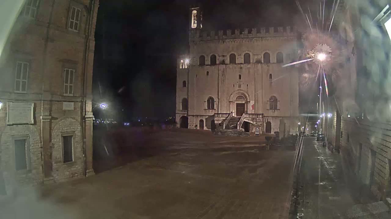A large, stone building with a staircase stands in a dark, empty square at night, fireworks exploding above it.