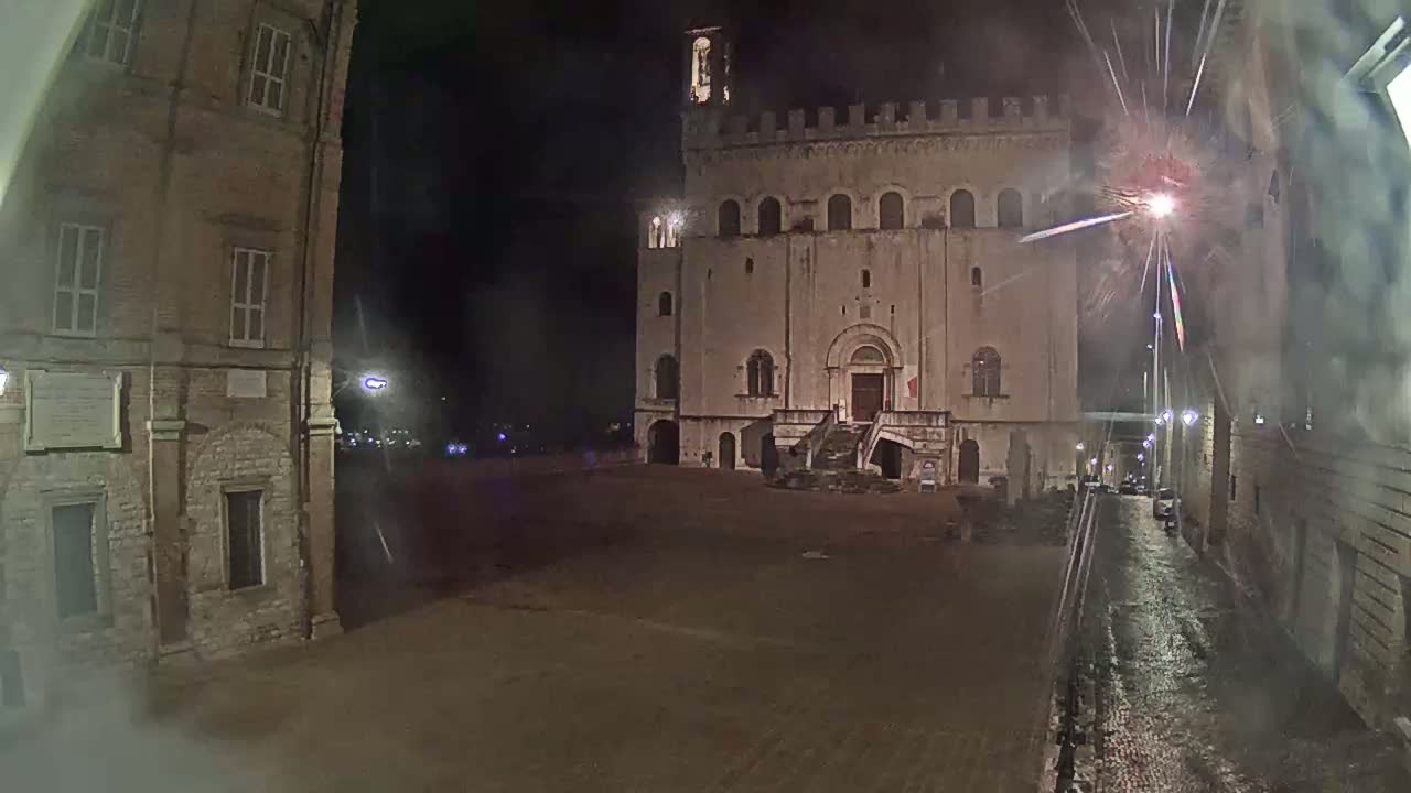 A large stone building stands in a plaza at night, with fireworks visible in the sky and rain or moisture on the ground.