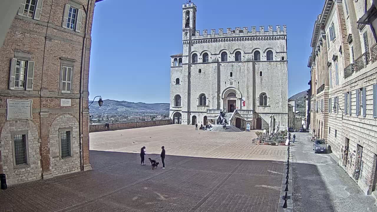 A large, light-colored stone building stands in a sunny town square, flanked by brick buildings, with a few people and dogs in the square and a car parked on a nearby street.