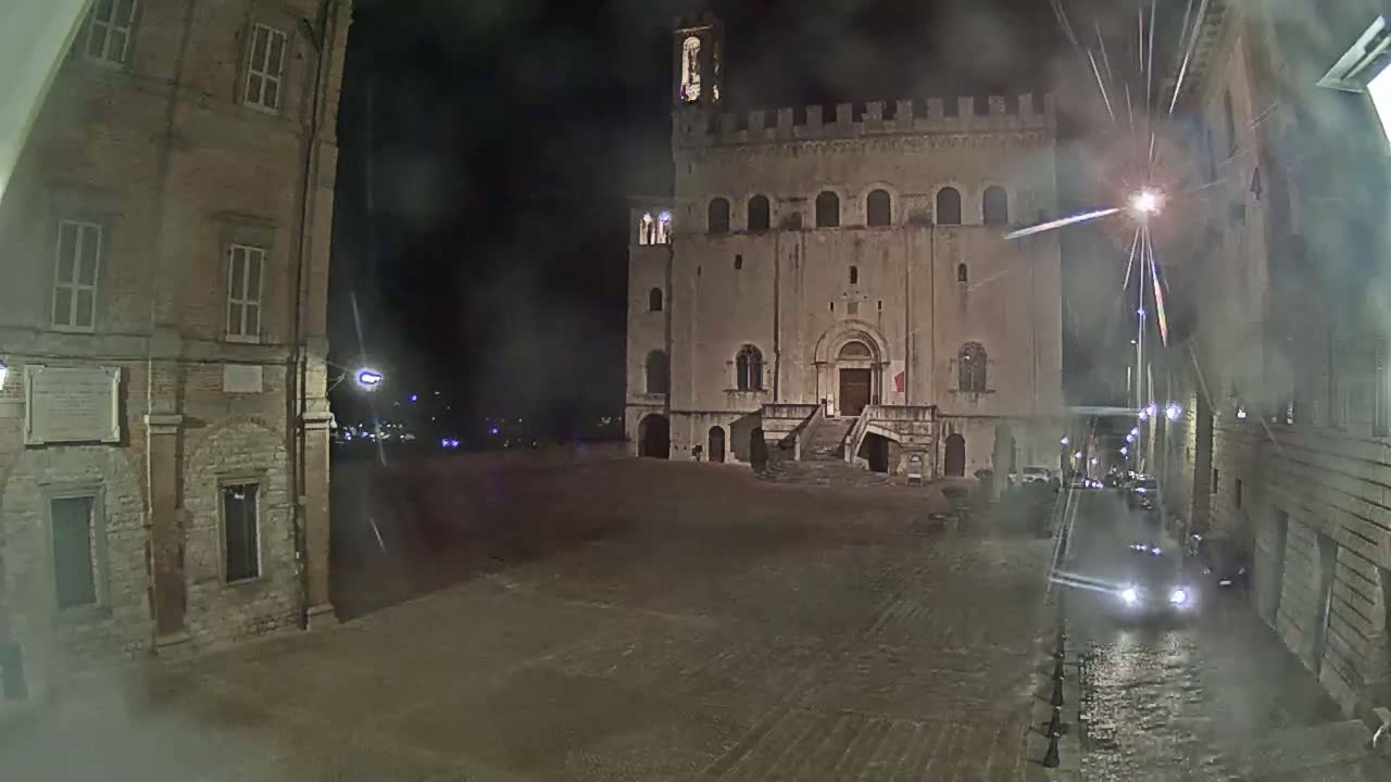 A large, stone building with a staircase stands in a dimly lit, empty square at night, under a misty or rainy sky.