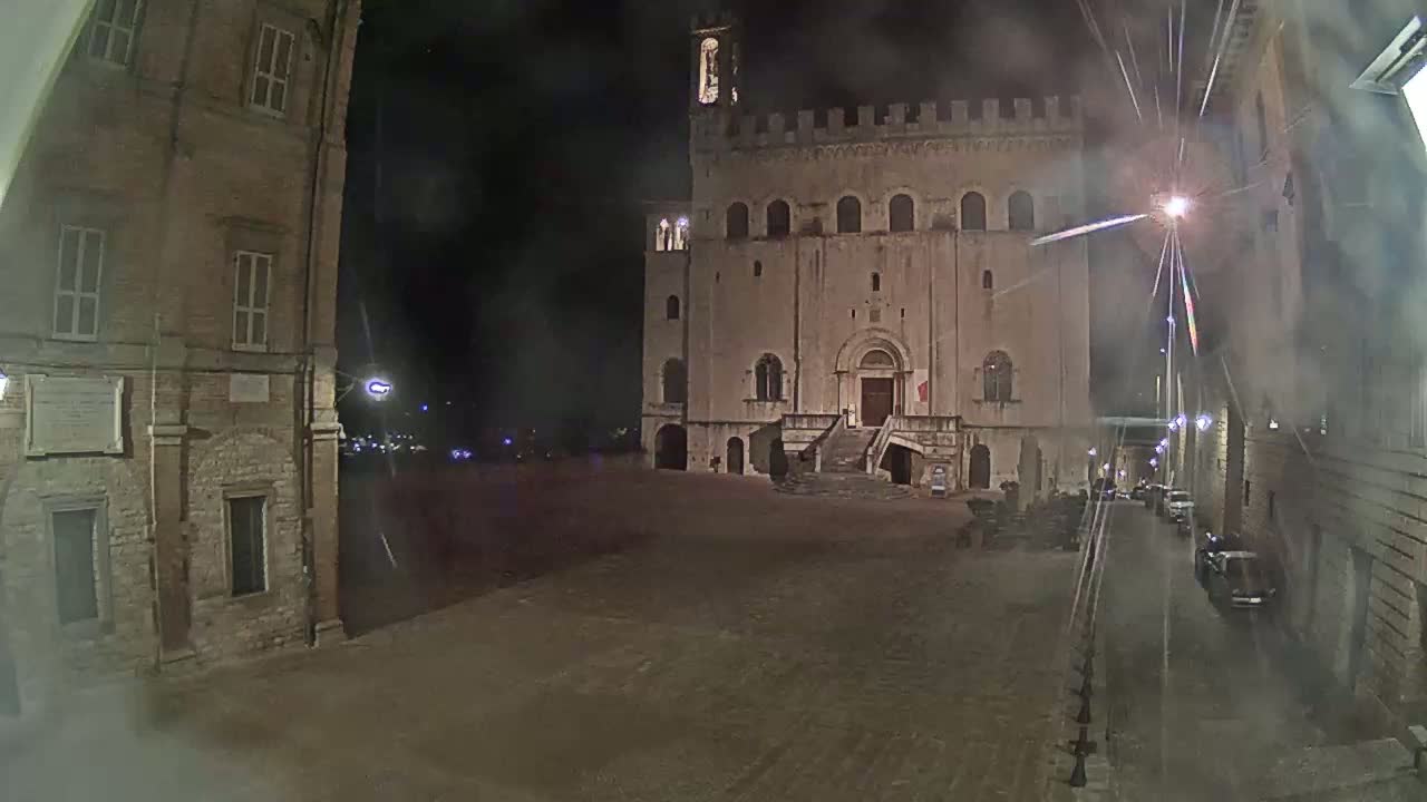 A large, stone building with a crenellated top stands in a mostly empty square at night, under hazy conditions.