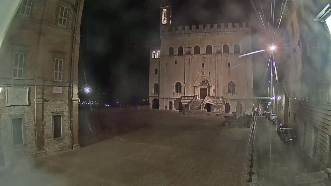 A large, stone building with a staircase stands in a dimly lit, empty plaza at night, flanked by other buildings and a street with a few parked cars.