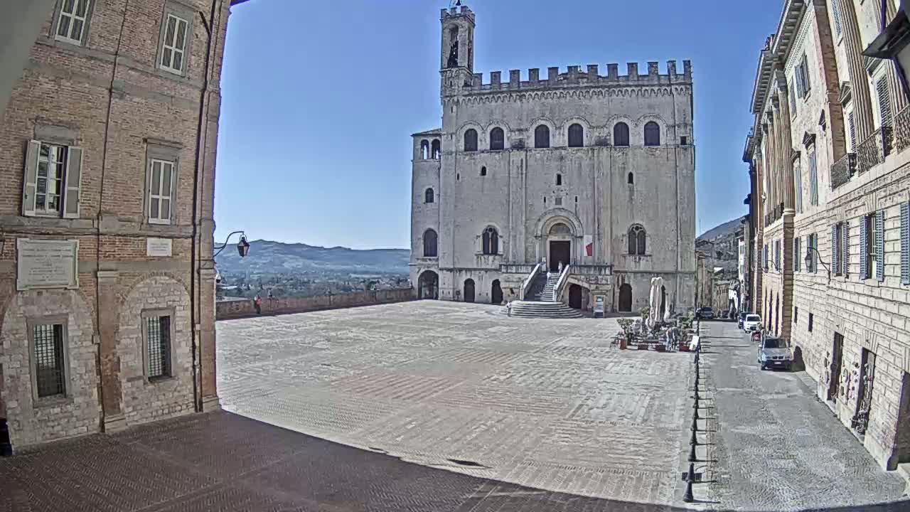 A large, stone public square is viewed from between two brick buildings on a sunny day, with a large, imposing stone building at the far end of the square.