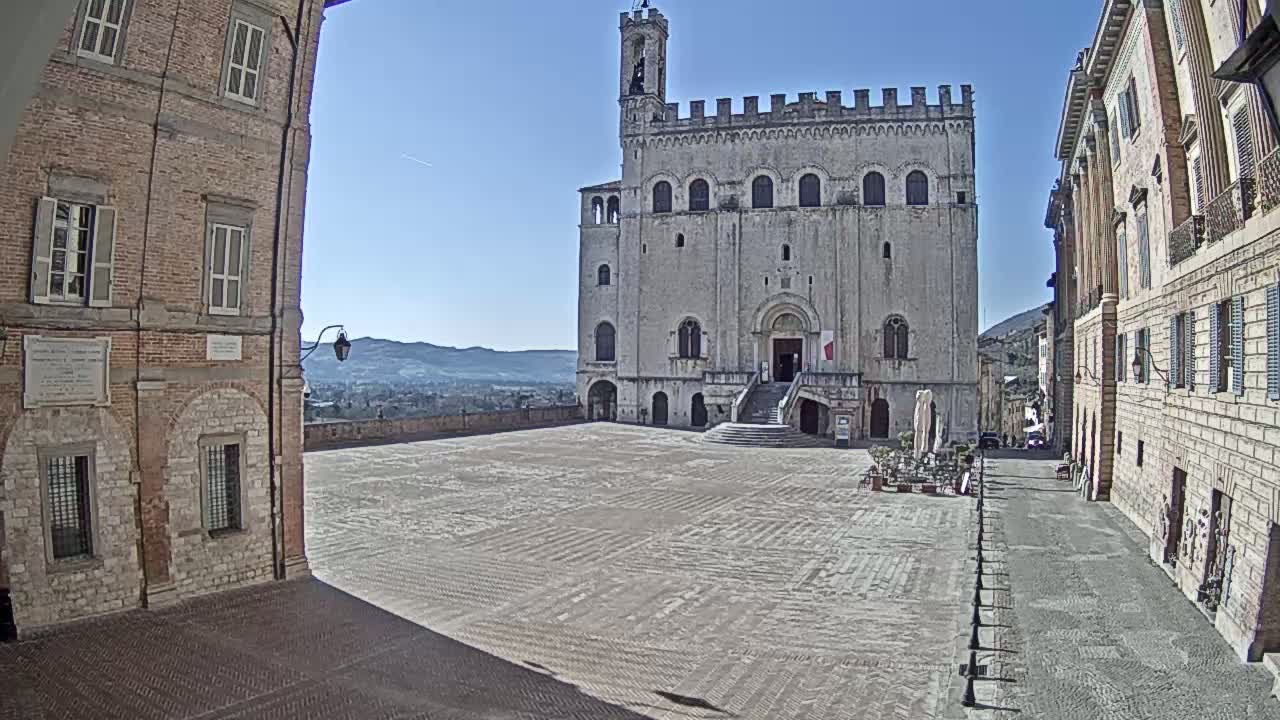 A large, light-colored stone building with a tower sits in a wide, empty plaza flanked by brick buildings, under a clear blue sky.