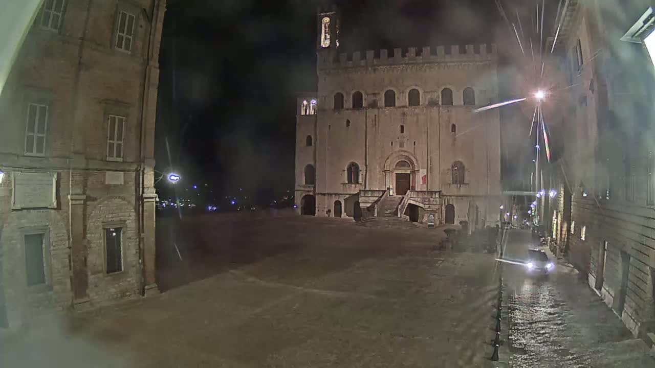 A large stone building with a staircase stands in a square at night in wet conditions, flanked by smaller stone buildings.