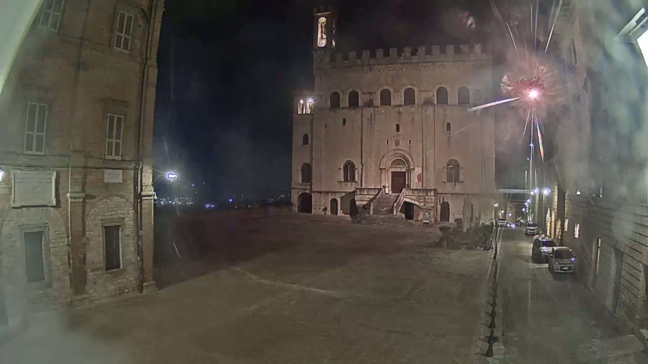 A large stone building with a staircase stands in a square at night, with fireworks exploding in the sky.