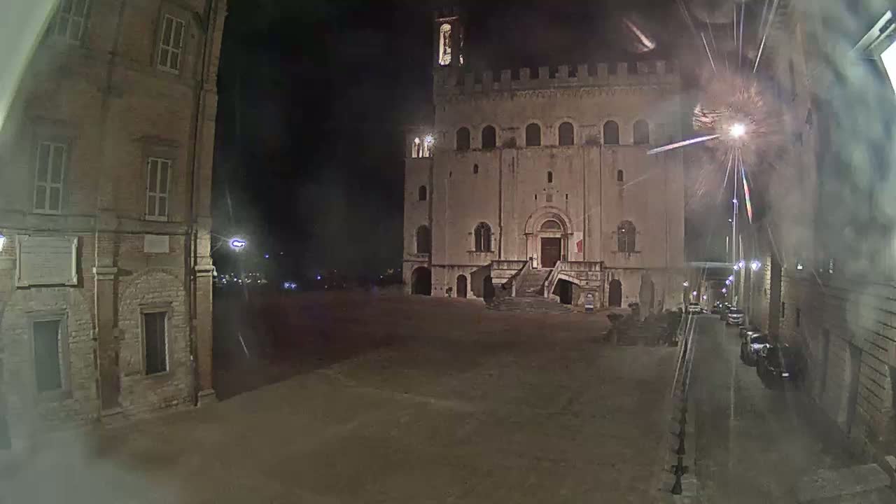 A large, stone building with a staircase stands in a mostly empty square at night, under a dark sky with faint light streaks.