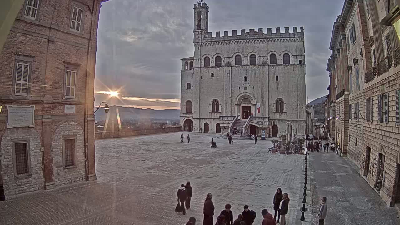 A large stone plaza is surrounded by old buildings, with a large pale stone building at the far end, and the setting sun visible in the distance under a cloudy sky.