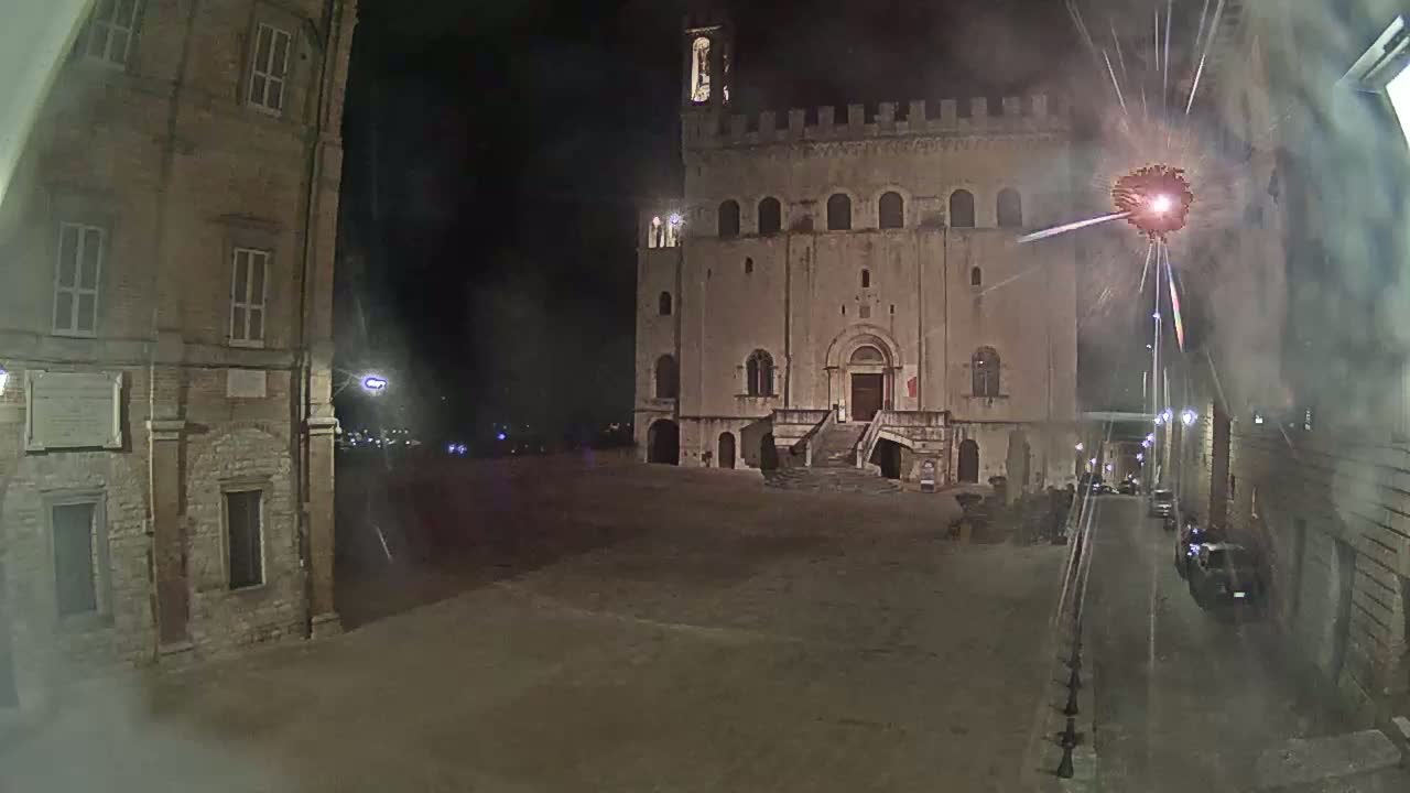A large stone building with a staircase stands in a town square at night, illuminated by lights and a firework display.