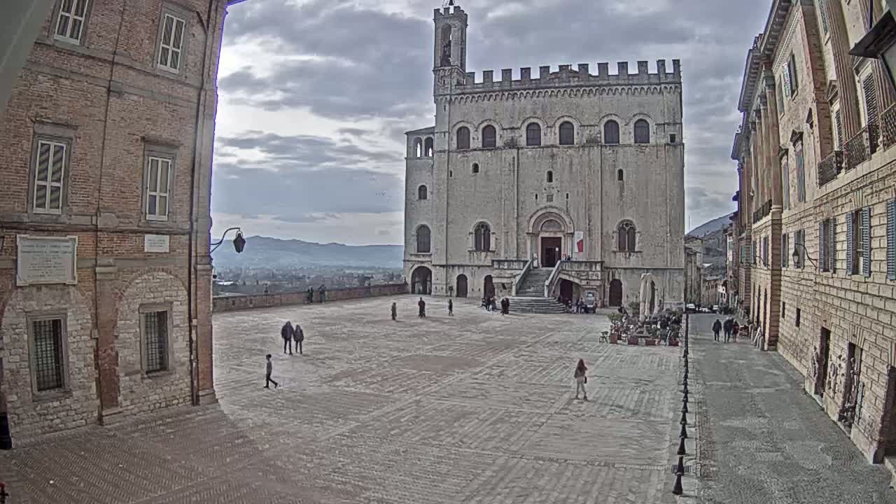 A large, stone plaza is surrounded by historic buildings under a cloudy sky, with a few people walking about.