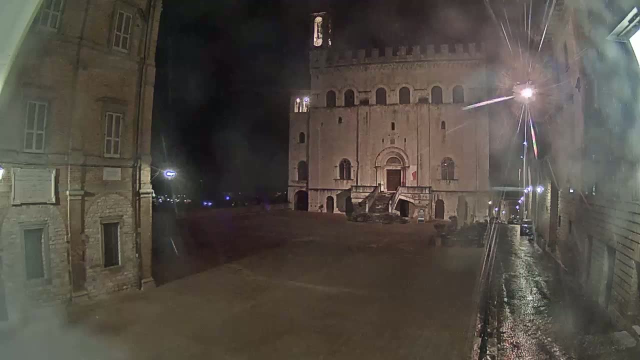 A large, stone building with a staircase stands in a square at night, under a rainy sky.