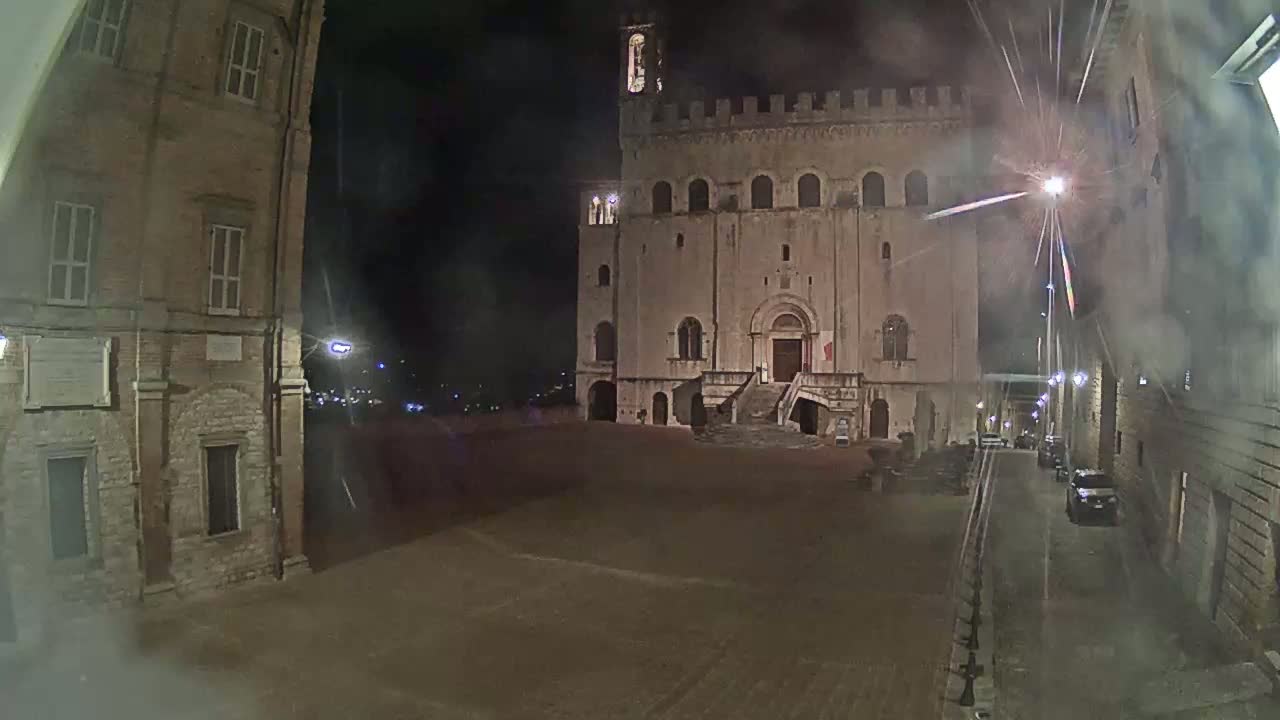 A large stone building with a staircase stands in a dimly lit, empty square at night, with hazy weather conditions and a few cars visible down a side street.