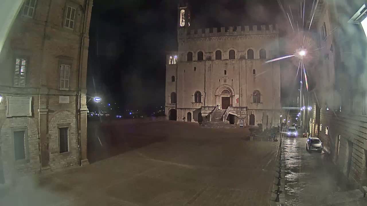 A large stone building with a staircase stands in a square at night in the rain, flanked by other buildings.