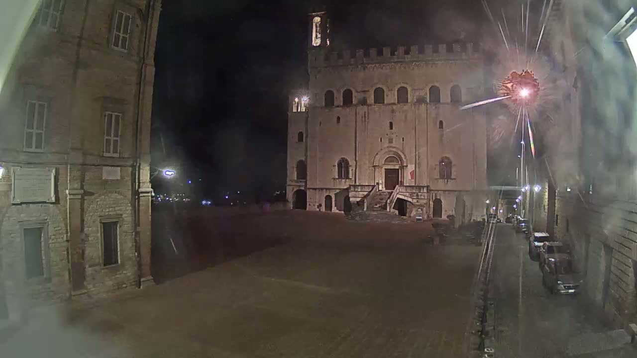 A large stone building with a staircase is seen at night in a mostly empty square, with fireworks exploding in the sky and several cars parked along a street.