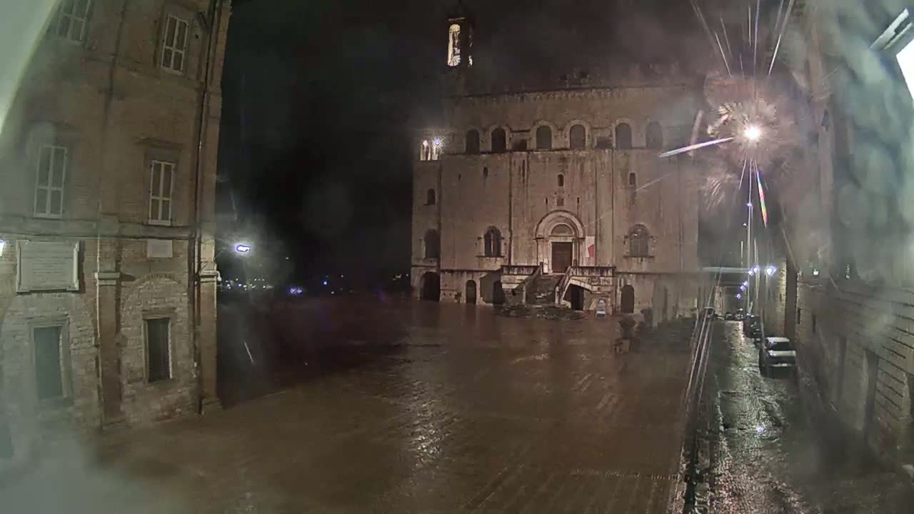 A flooded, cobblestone town square at night, featuring an old stone building with a stairway, lit by streetlights under fireworks.