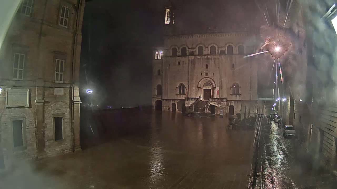 A large stone building stands in a flooded square at night during a heavy rainstorm, with fireworks exploding in the distance.