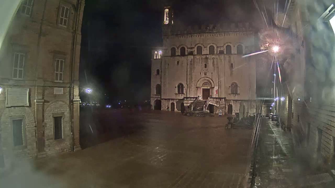 A large, stone building stands in a dimly lit square at night under a rain shower.