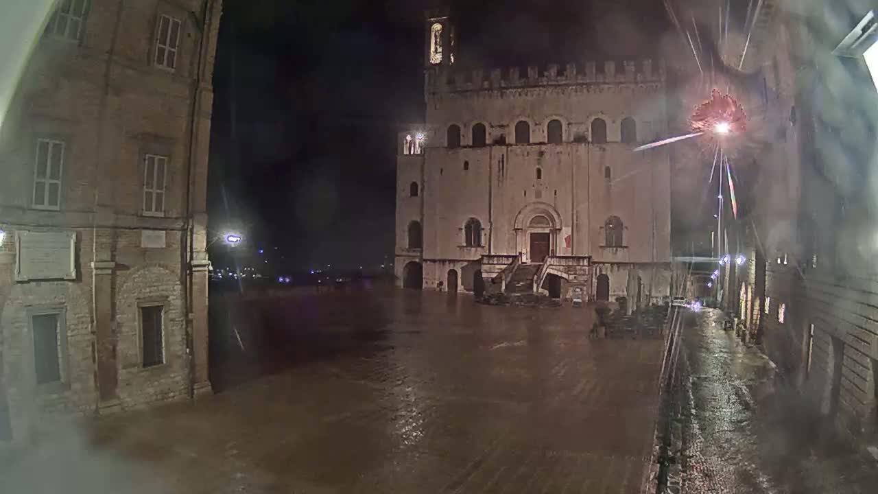 A large, stone building stands in a floodlit town square at night during a rain storm, with fireworks exploding in the background.