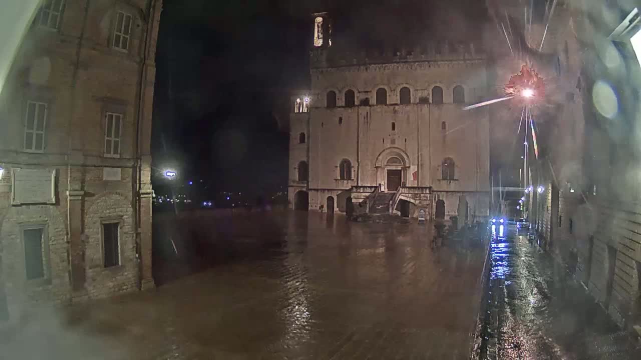 Floodwaters at night engulf a town square and street, with old stone buildings rising from the submerged area under heavy rain.