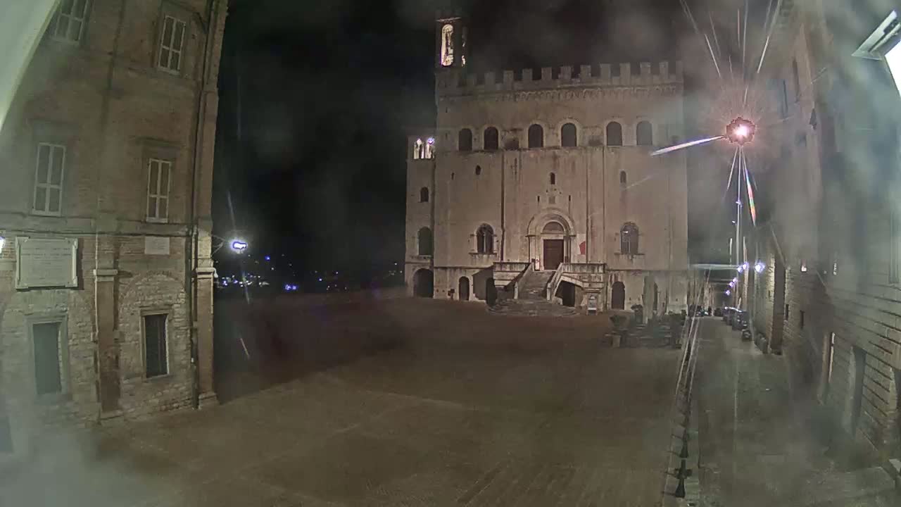 A large, stone building with a staircase stands in a mostly empty square at night under a misty sky.