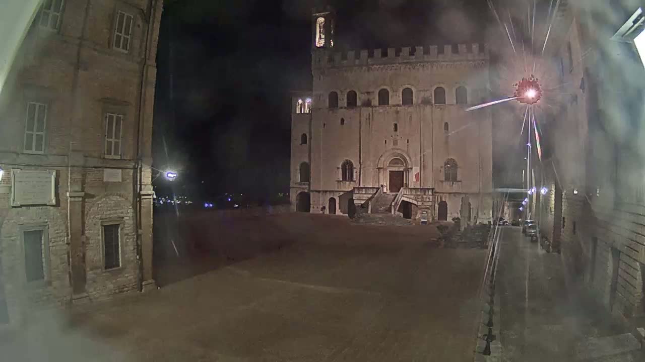 A large, stone building with a staircase stands in a dimly lit square at night, with fireworks visible in the sky.