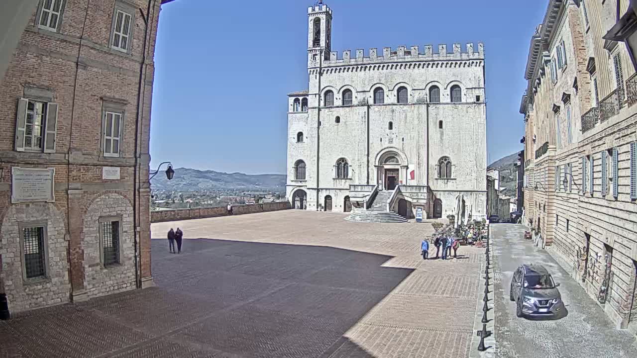 A large, light-colored stone building with a tower stands in a brick plaza on a sunny day, with brick buildings lining the sides and distant hills visible in the background.