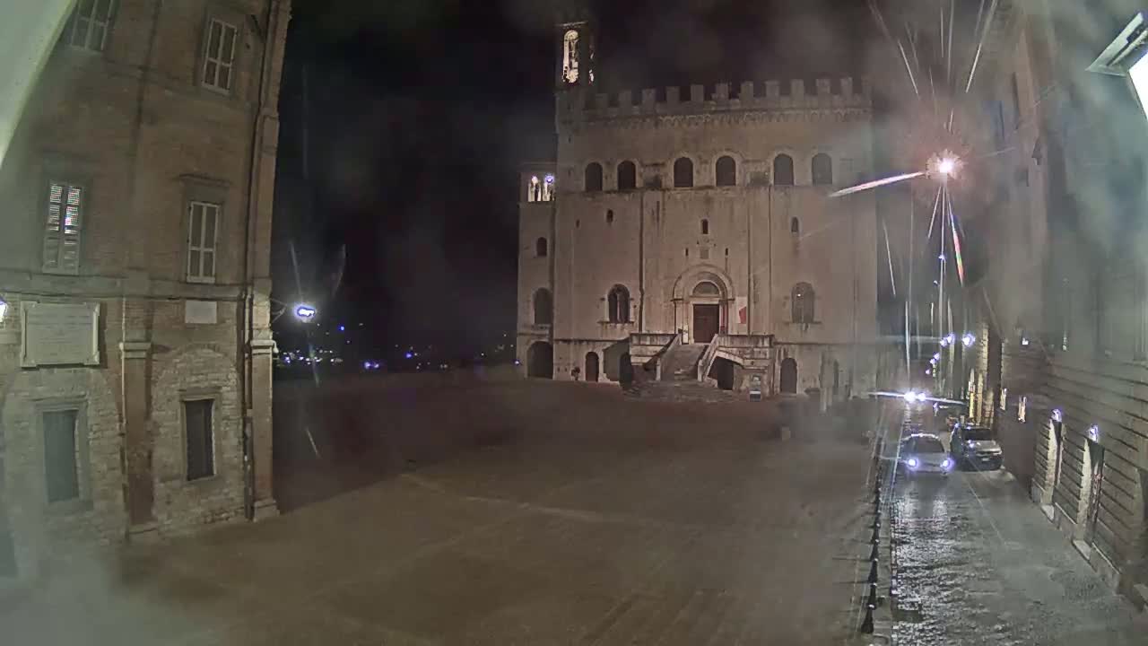 A large, stone building with a staircase stands in a mostly empty square at night, under a dark, possibly rainy sky.