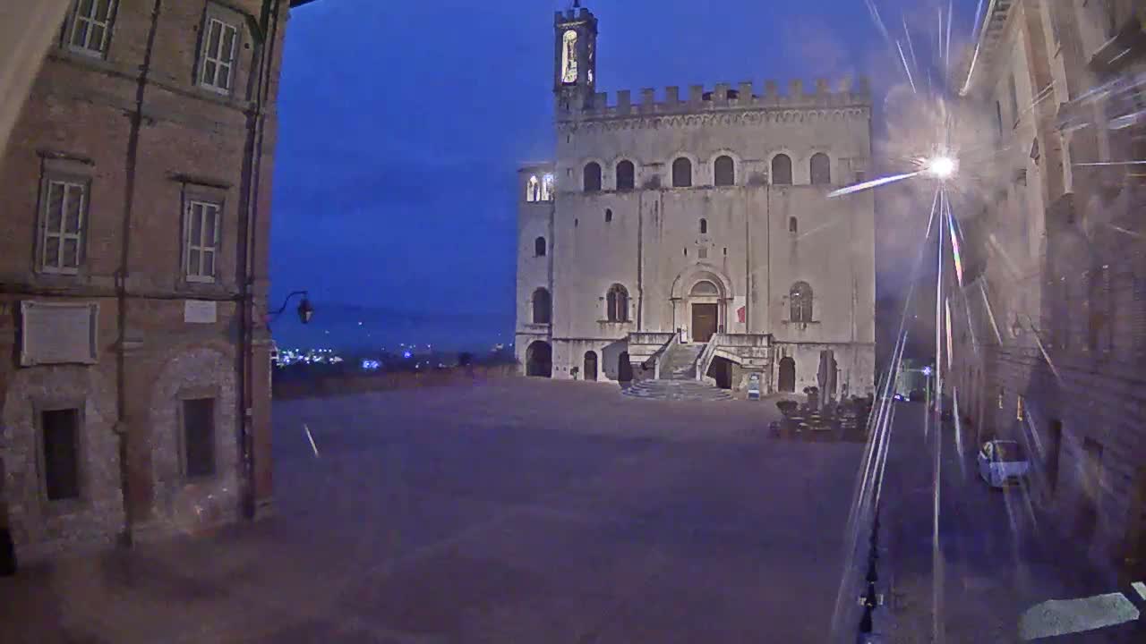 A large, pale stone building with a tower stands in a square at dusk, flanked by older brick buildings under a dark blue, possibly stormy sky.