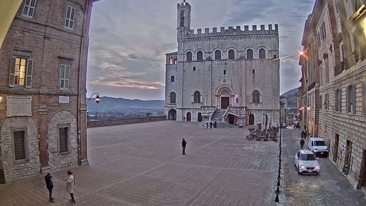 A large, light-colored stone building with a tower stands in a town square under a dusky sky, with a few people visible in the plaza and cars parked along a nearby street.
