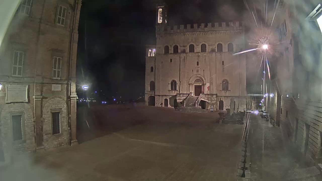 A large, stone building with a staircase stands in a dimly lit, empty square at night, under hazy conditions.