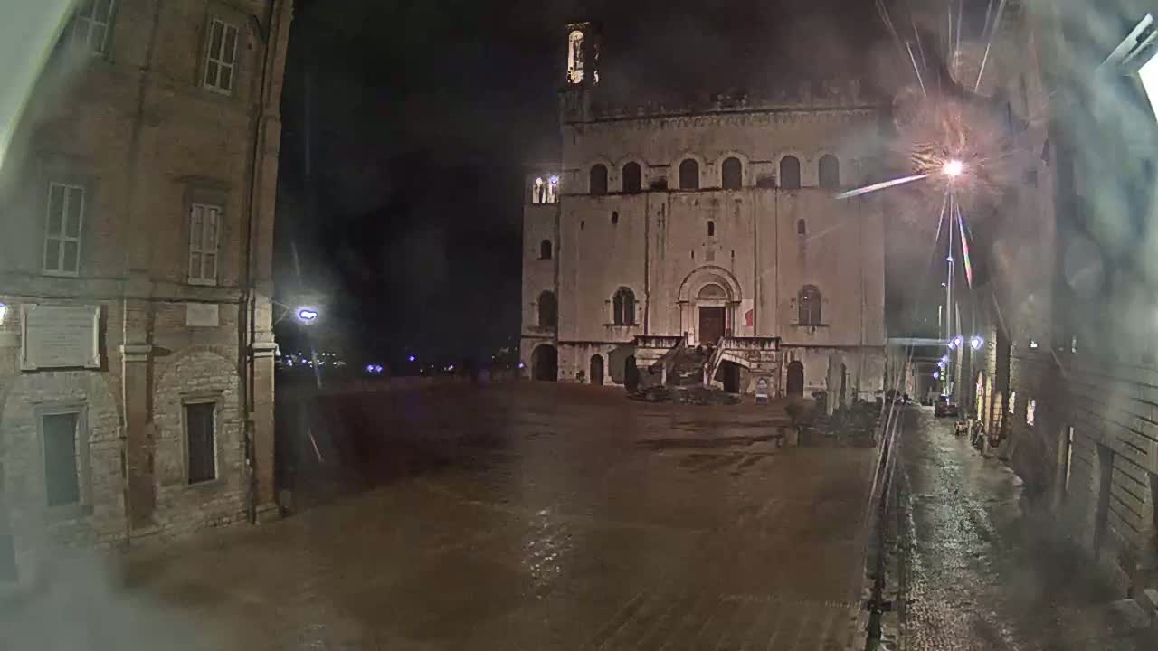 A large, stone building stands in a square at night under a rainy sky, flanked by smaller buildings, with wet cobblestones visible throughout.