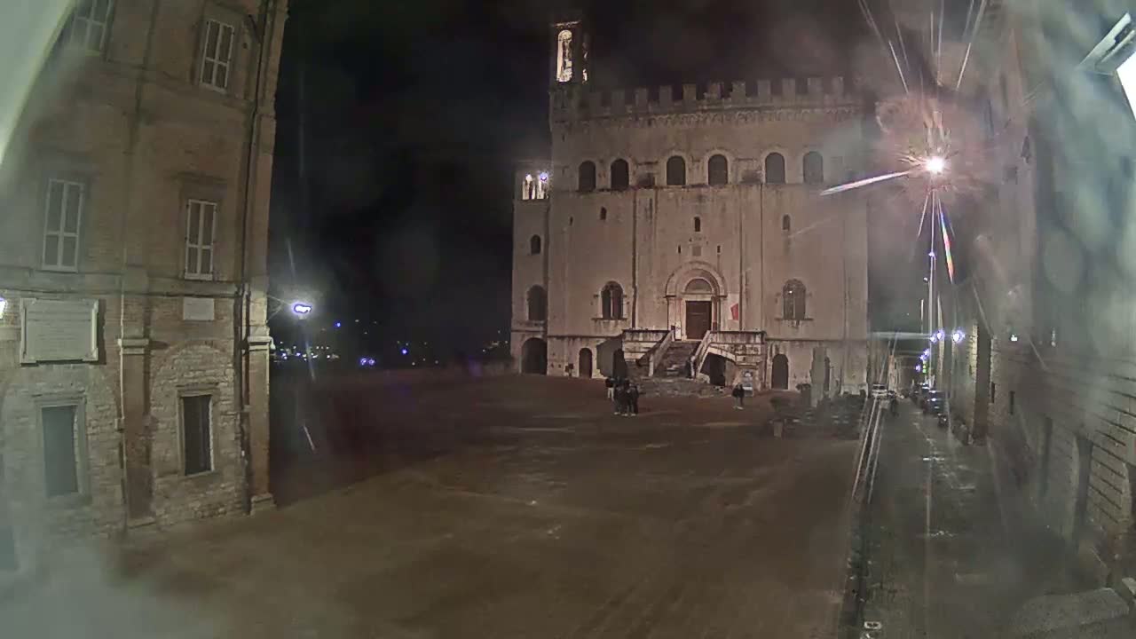 A large, pale stone building with a staircase stands in a plaza at night, flanked by older buildings, under a dark, possibly rainy sky with firework-like streaks of light visible in the air.