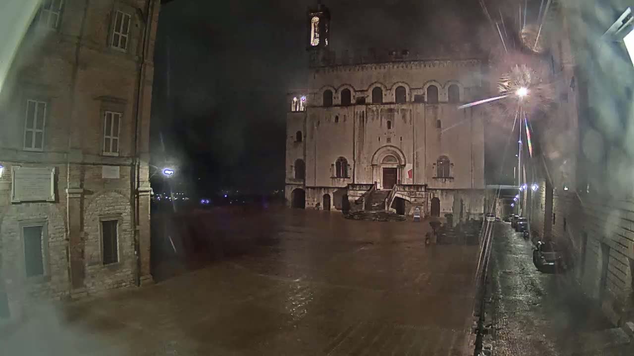 A large, flood-covered square at night is flanked by ancient stone buildings under a rainy sky.