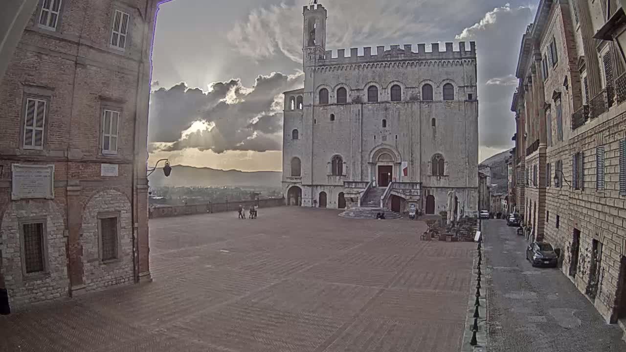 A large, light-colored stone building with a staircase stands in a town square under a partly cloudy sky, with the sun breaking through the clouds in the distance.