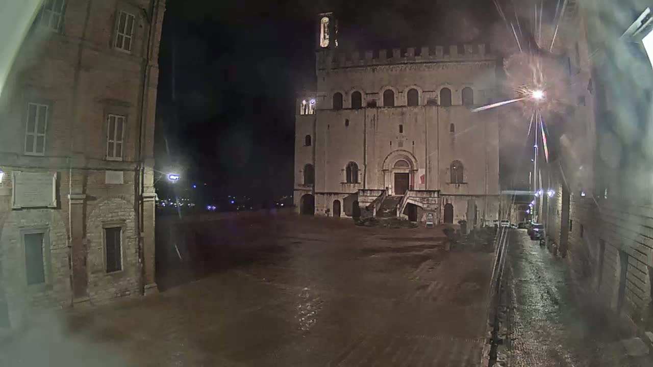 A large, stone building with a staircase stands in a wet, empty square at night, flanked by smaller buildings and a narrow street.