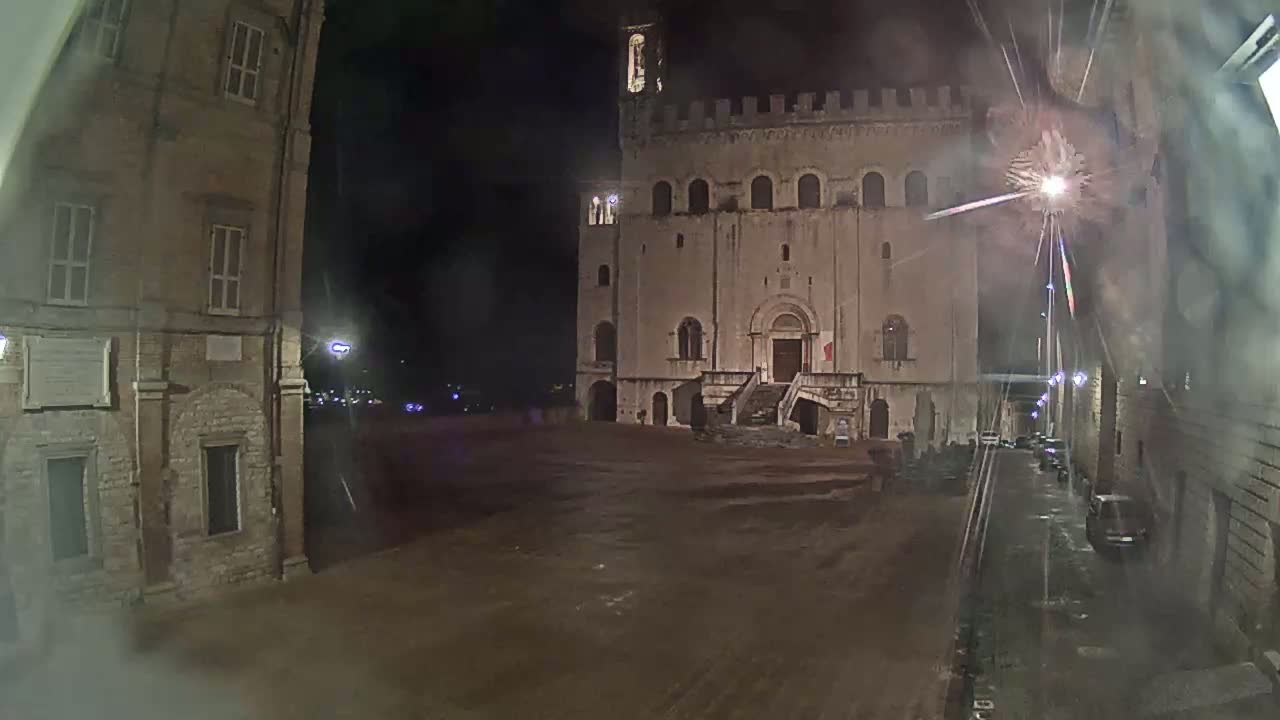 A large, stone building with a staircase stands in a dark, empty square at night, with fireworks visible in the sky.