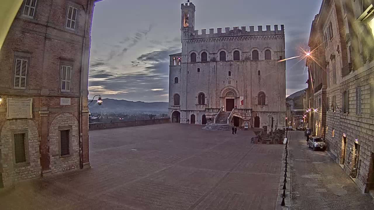 A large, light-colored stone building with a tower stands in a town square at dusk, under a cloudy sky.