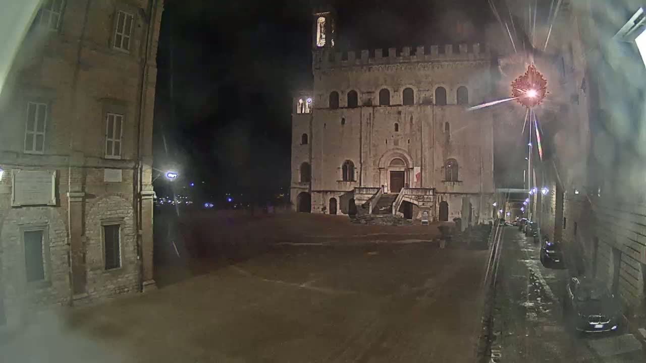 A large stone building with a staircase and tower is visible at night, with a few cars parked in a nearby street under dim lighting.