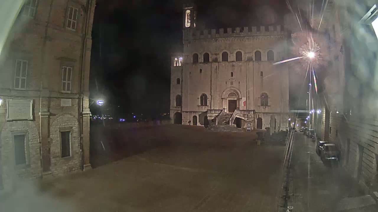 A large, stone building with a staircase stands in a square at night, with a few cars visible in a nearby street under a dark, possibly rainy sky.