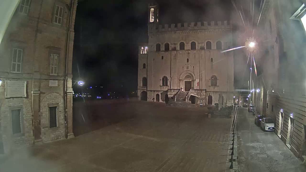 A large, stone building with a staircase stands in a plaza at night under hazy conditions, with a few cars visible on a nearby street.