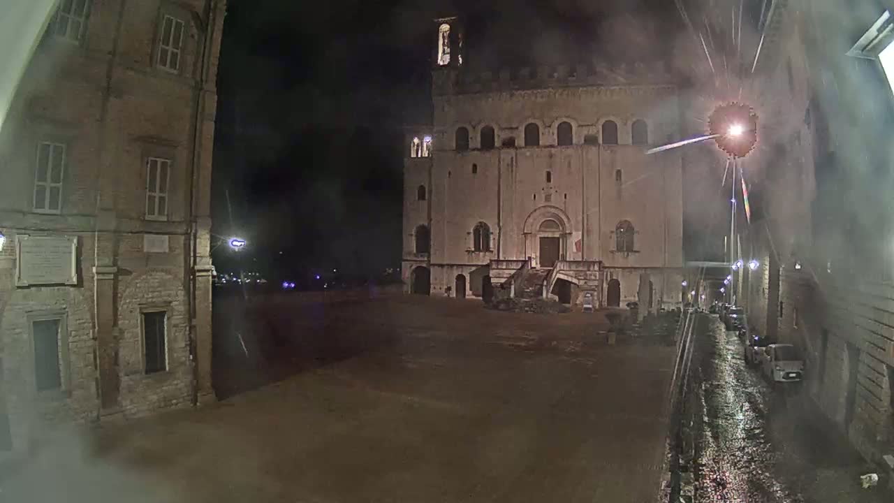 At night, under a fireworks display, a large, tan stone building with a staircase stands in a wet, empty plaza between two other buildings.