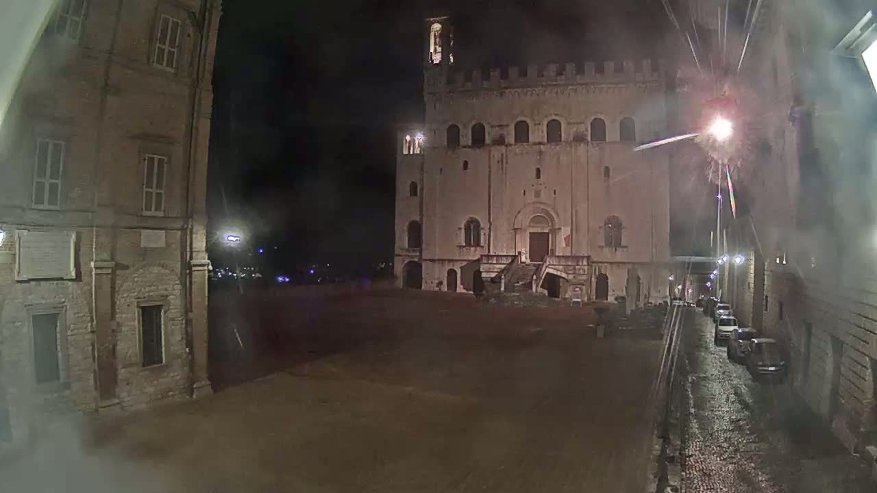A large, tan stone building with steps leading to its entrance sits in a dimly lit plaza at night, with a street and parked cars visible to one side.