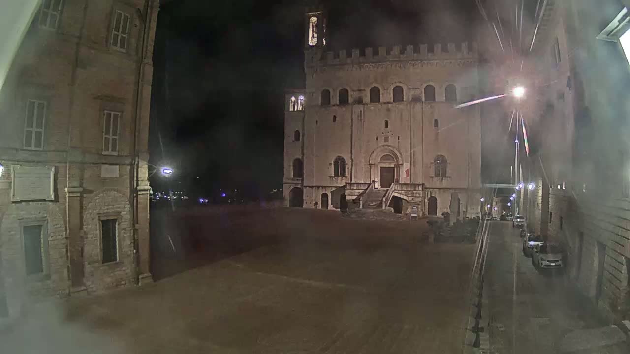 A large, stone building stands in a dimly lit square at night, with a few cars parked along a narrow street to the right.