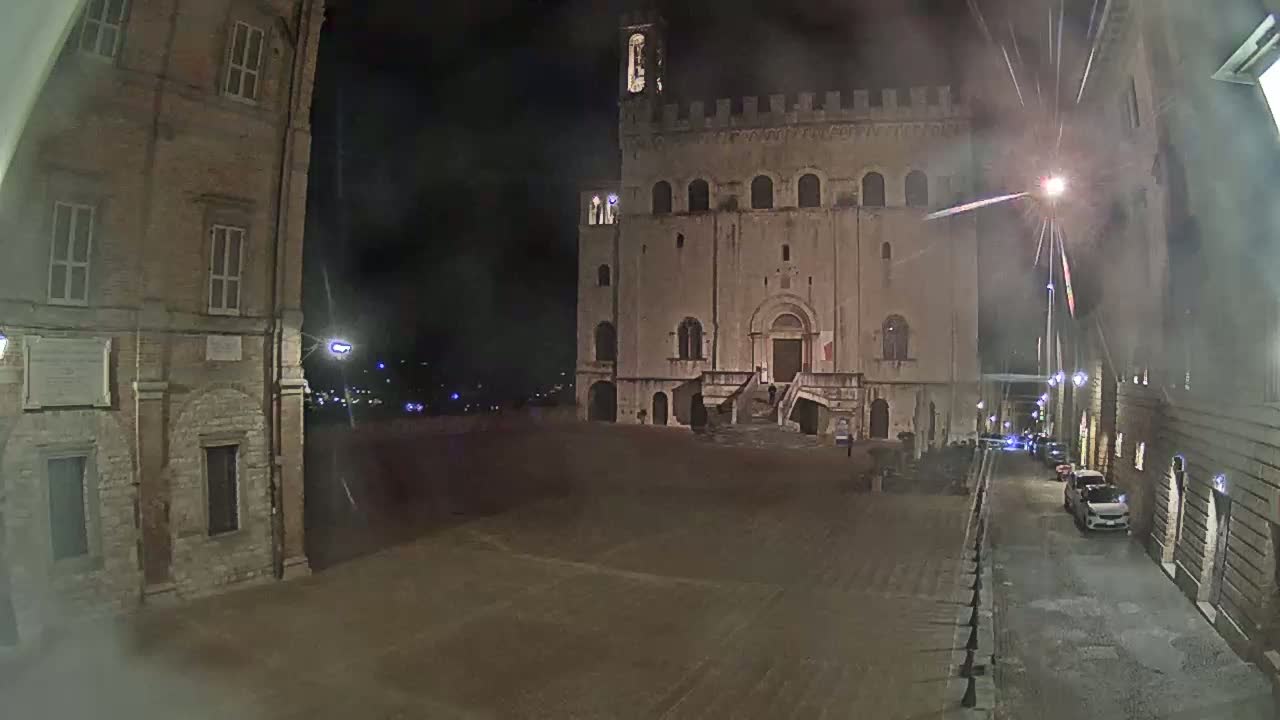 A large, stone building with a tower stands in a dark, empty plaza at night, flanked by other buildings and a few parked cars.
