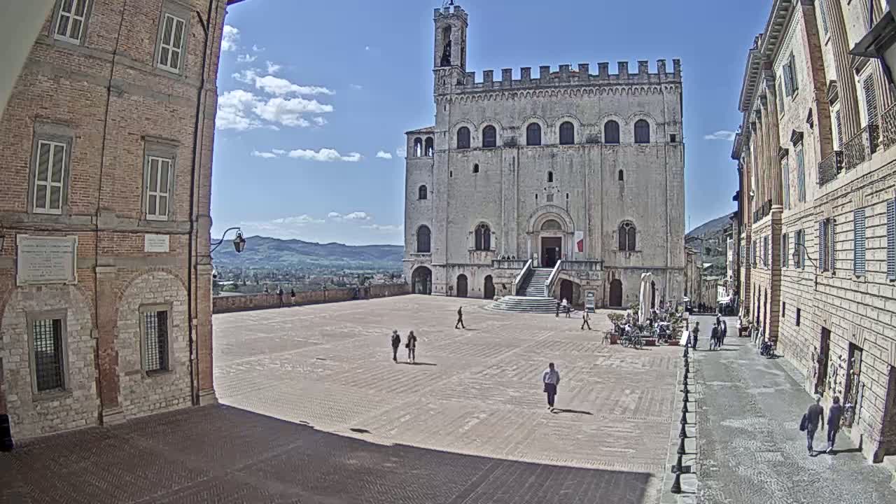 A large, light-colored stone building with a tower stands in a town square on a sunny day, with people walking in the square and along the adjacent streets.