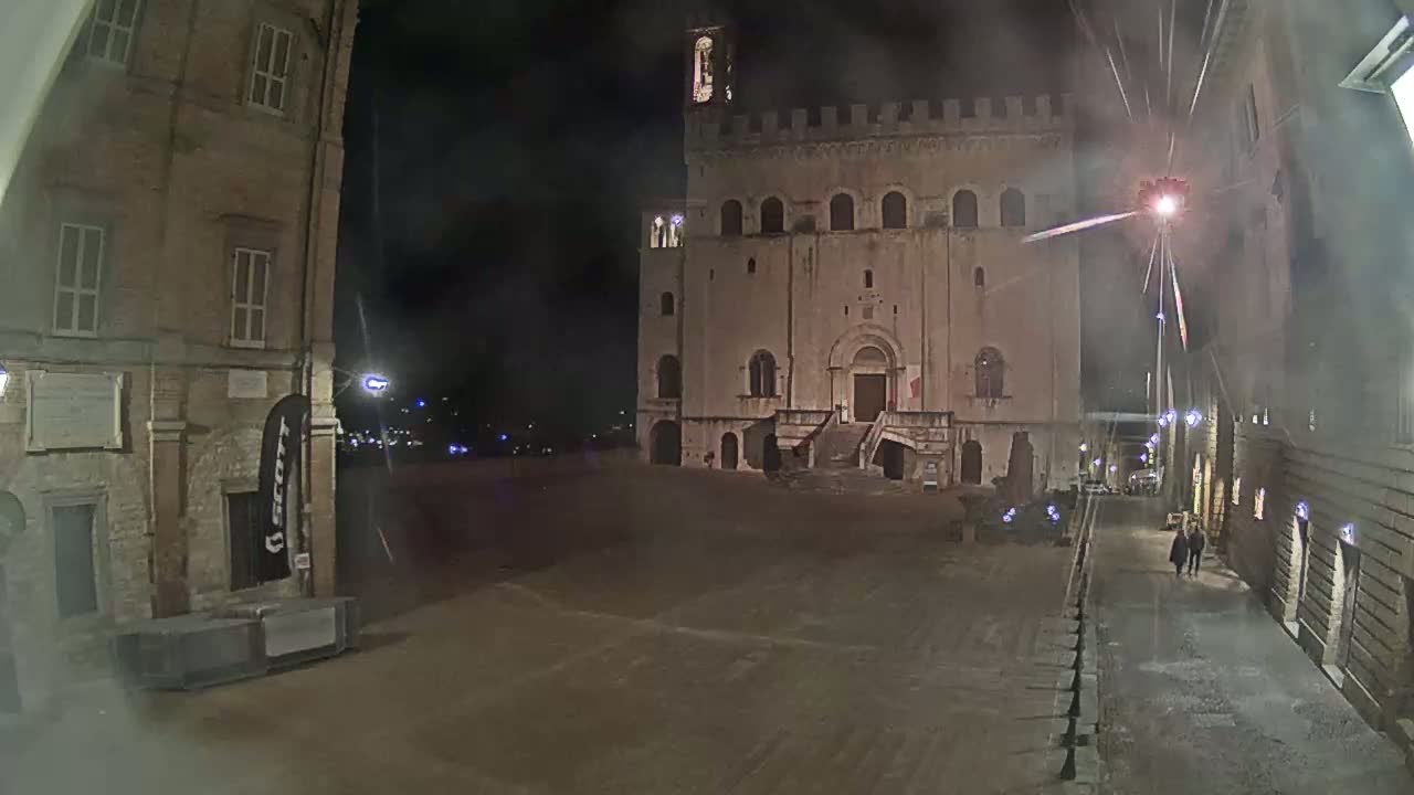 A large, stone building with a staircase stands in a mostly empty, dimly lit square at night, with a few people walking along a nearby street.