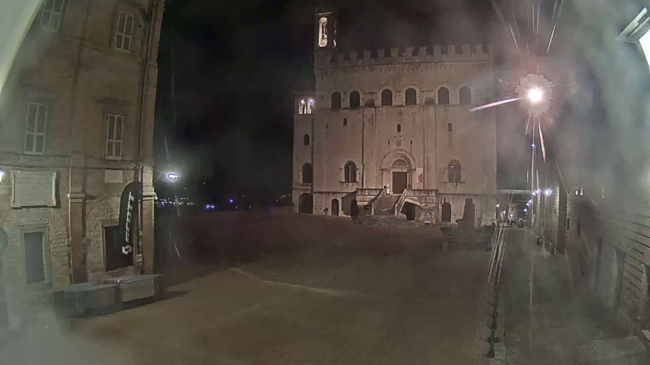 A large, stone building with a staircase stands in a mostly empty square at night, under hazy or smoky conditions.